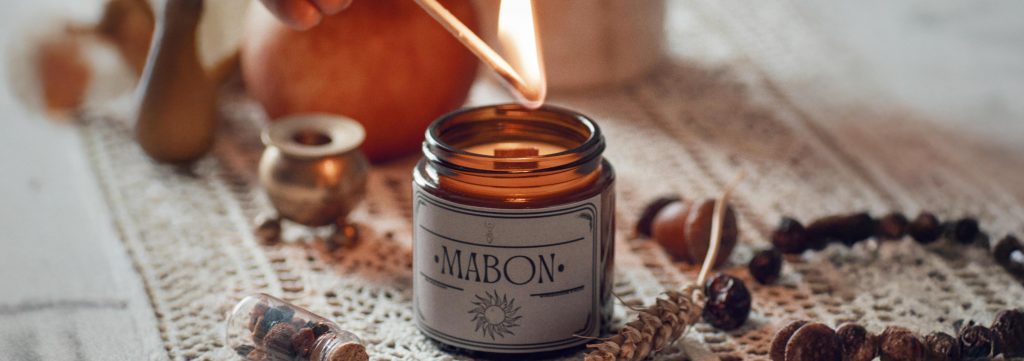A lit candle labelled “Mabon” surrounded by acorns, beads, and autumn harvest decorations.