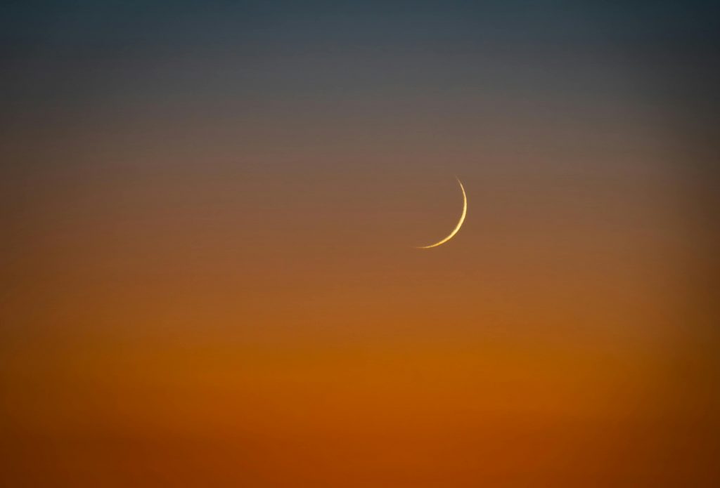 Thin crescent new moon in a glowing orange sunset sky